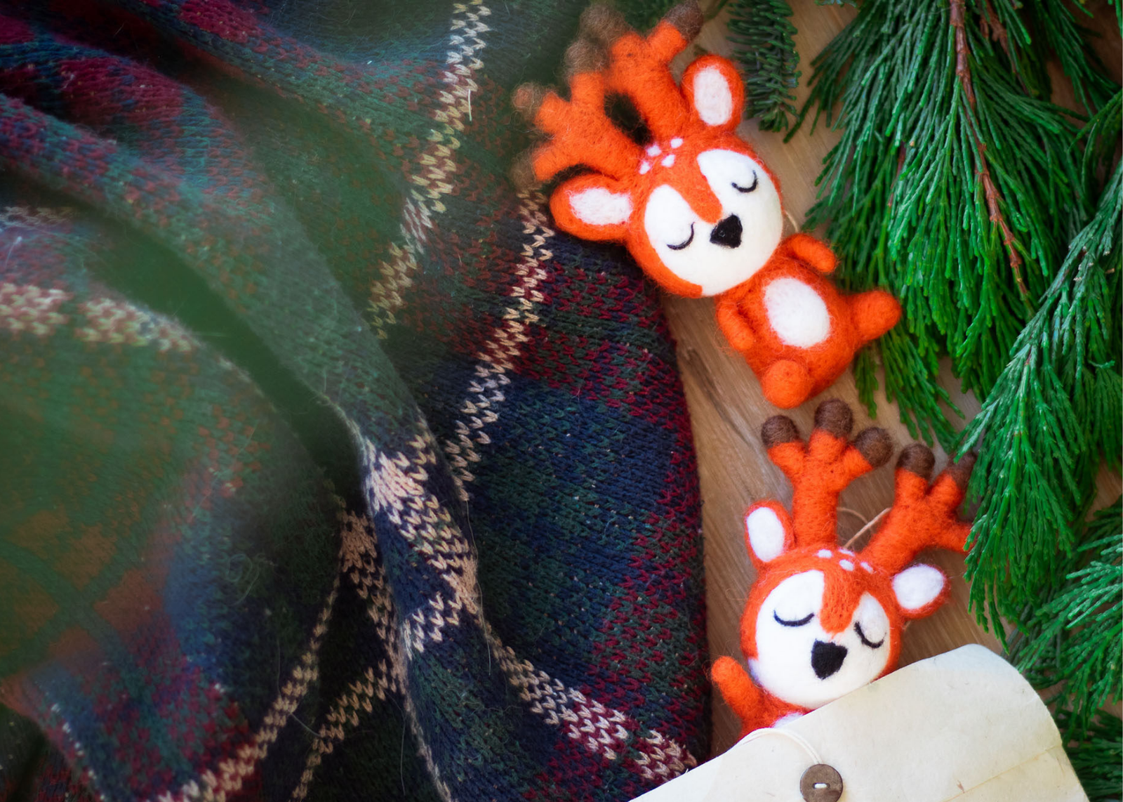 Two orange wool deer lay on a plaid holiday sweater next to spruce branches.