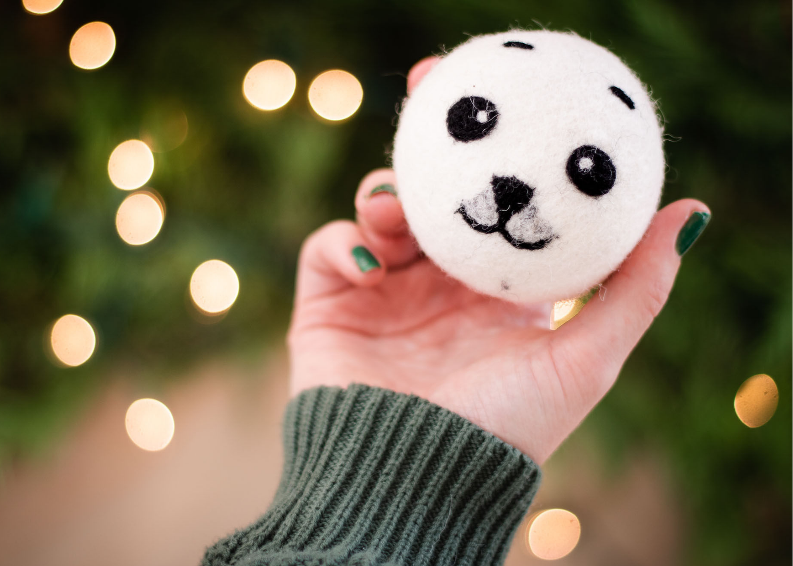 a hand is holding a white dryer ball in a cute black baby seal design, yellow holiday lights are on the dark green background
