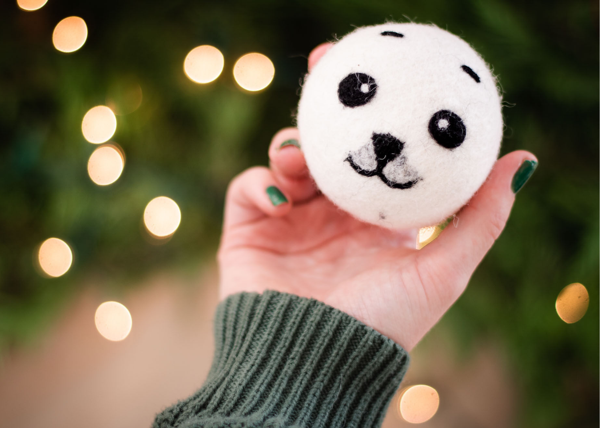  a hand is holding a white dryer ball in a cute black baby seal design, yellow holiday lights are on the dark green background