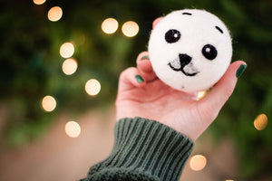  a hand is holding a white dryer ball in a cute black baby seal design, yellow holiday lights are on the dark green background