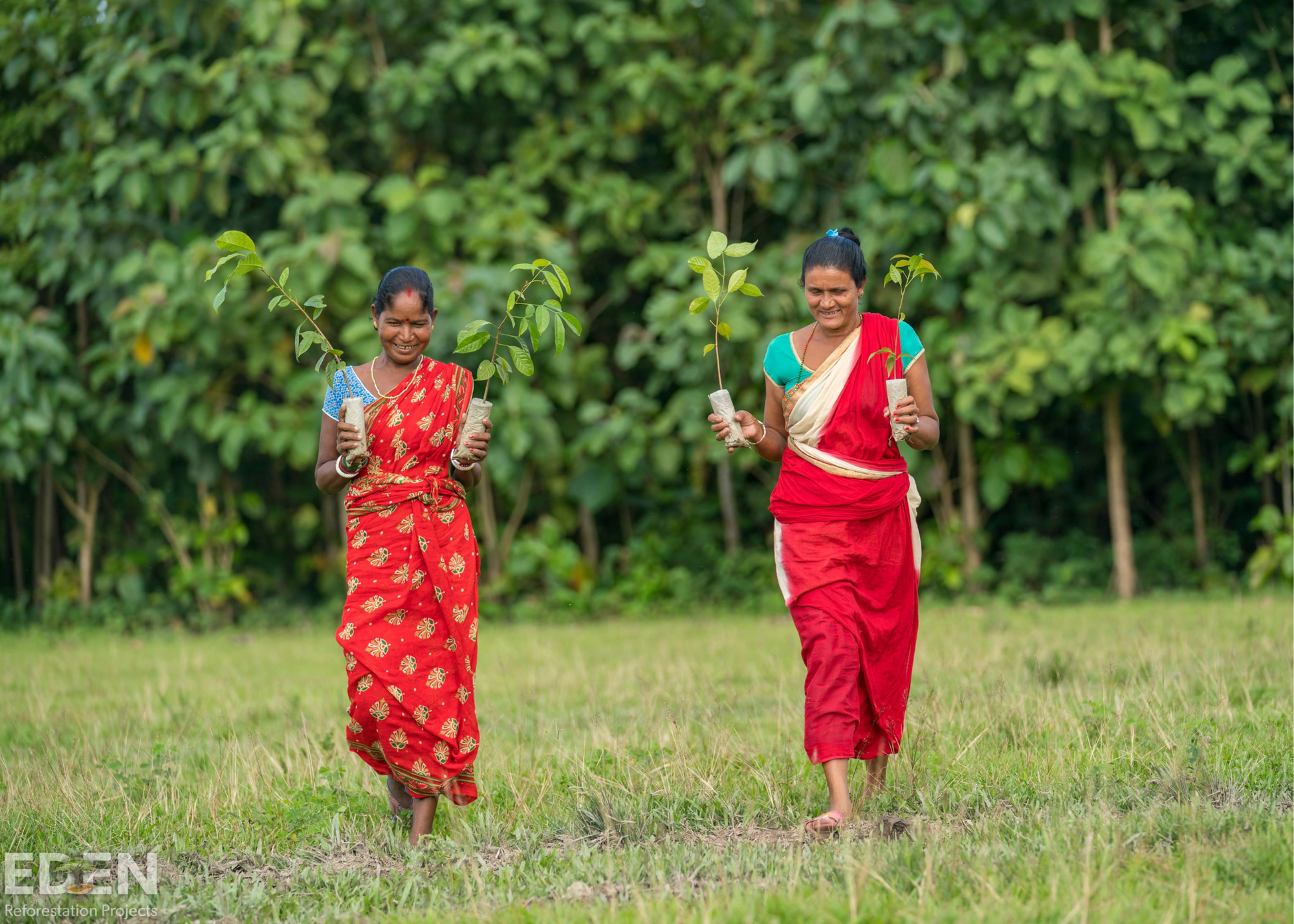 two smiling women walk in the grass holding plants in each hand