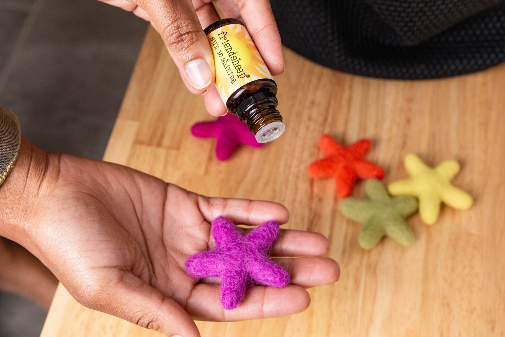 A violet star freshener is in a hand. The other hand is dropping some essential oil on it. On the background 4 more colorful star fresheners